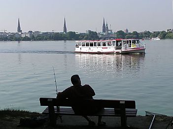 011_25828 | Hamburg Panorama mit den Kirchtrmen der Hauptkirchen; der Alsterdampfer fhrt auf der Aussenalster Richtung Winterhude - ein Angler sitzt auf einer Bank und beobachtet die Szene.