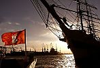 011_25991 |  Sonnenuntergang im Hamburger Hafen; an den St. Pauli Landungsbrcken liegt ein Grosssegler - im Hintergrund am gegenber liegenden Elbufer die Werft Blohm + Voss, rechts weht die Hamburg Fahne im Wind.