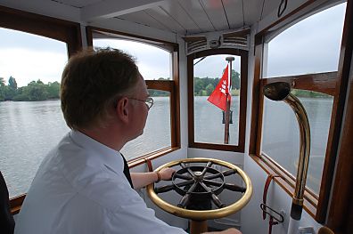 011_25949 | der Conducteur des historischen Alsterschiffs in seiner Fahrerkabine mit der Hand am Messing Steuerrad - rechts das Mundstck der Sprechanlage; am Bug des Schiffs die Hamburg Fahne.
