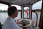 011_25949 | der Conducteur des historischen Alsterschiffs in seiner Fahrerkabine mit der Hand am Messing Steuerrad - rechts das Mundstck der Sprechanlage; am Bug des Schiffs die Hamburg Fahne.