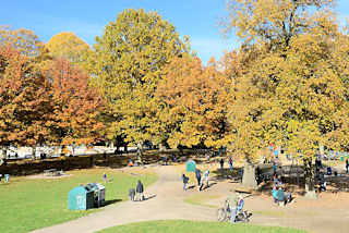 6646 Blick über den Kinderspielplatz beim Planschbecken im Hamburger Stadtpark - hohe Bäume mit bunt gefärbtem Herbstlaub säumen die Wege und den Platz; Kinder spielen in der Sonne. 