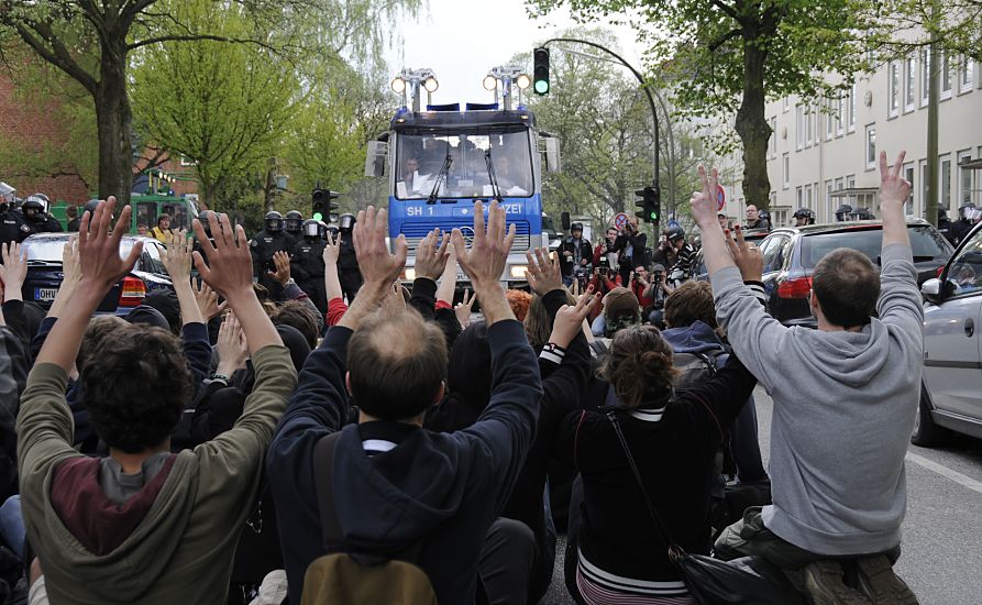 011_26019 |  Demonstranten haben sich auf dem Rbenkamp auf die Fahrbahn gesetzt und versuchen mit einer gewaltfreien Strassenblockade den Marsch der Neonazis durch den Hamburger Stadtteil Barmbek aufzuhalten - sie halten die Hnde hoch, um ihre Gewaltfreiheit zu demonstrieren. Ein Wasserwerfer ist aufgefahren, die Blockierer werden per Lautspreicher aufgefordert, die Strasse zu rumen. Links stehen Polizisten mit Helm, zur Rumung bereit - auf der rechten Seite halten Fotografen die Szene mit ihrer Kamera fest. www.fotograf-hamburg.de