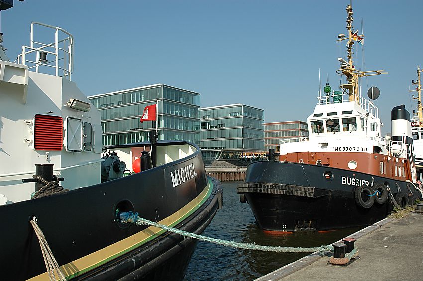 011_26031 |  Bilder aus Hamburg - Fotos vom Hamburger Hafen -- Am Bug vom Schlepper MICHEL weht die Hamburg Fahne im Wind; das Schiff ist mit starken Tauen am Anleger festgemacht. Daneben liegt ein BUGSIER-Schiff, der Bug ist mit dickem Gummi gepolstert - auch so kann das ca. 3000 PS starke Arbeitsboot Schiffe in Position bringen. Im Hintergrund die moderne Glasarchitektur an der Uferpromenade von Hamburg Neumhlen.  www.christoph-bellin.de 